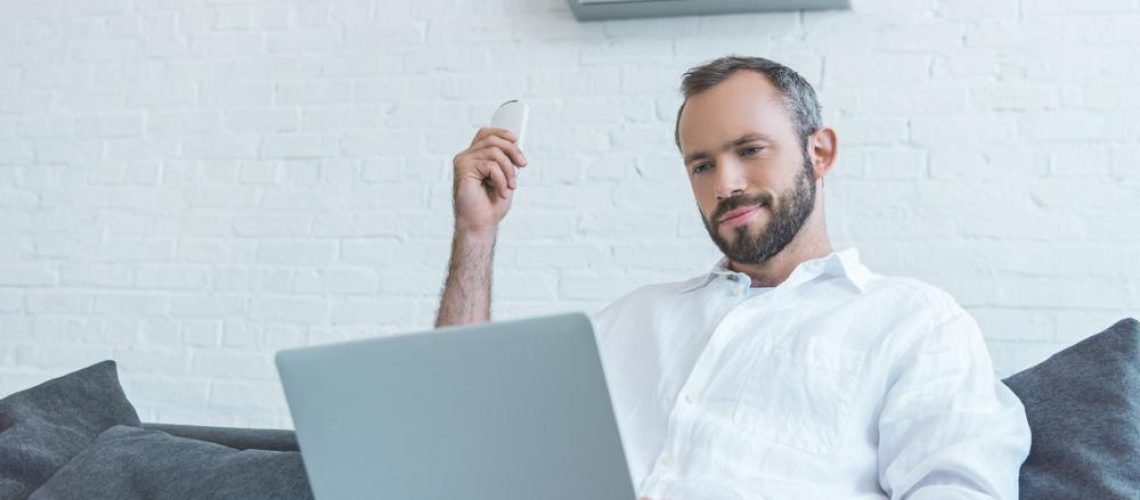 bearded man turning on air conditioner with remote control while using laptop